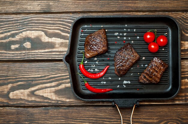 Gegrilltes Rindersteak in einer Grillpfanne mit Gewürzen, Tomaten, rotem Pfeffer auf Holzhintergrund