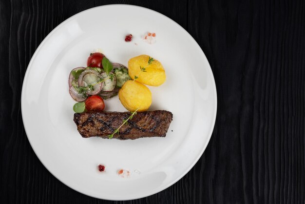 Gegrilltes Rindersteakfleisch mit Kartoffeln und Gemüse auf weißem Teller auf schwarzem Holzhintergrund wooden