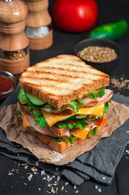 Gegrilltes Sandwich mit Schinken, Käse und frischem Gemüse auf dunklem Hintergrund mit Zutaten und Gewürzen. Vertikale Ansicht.