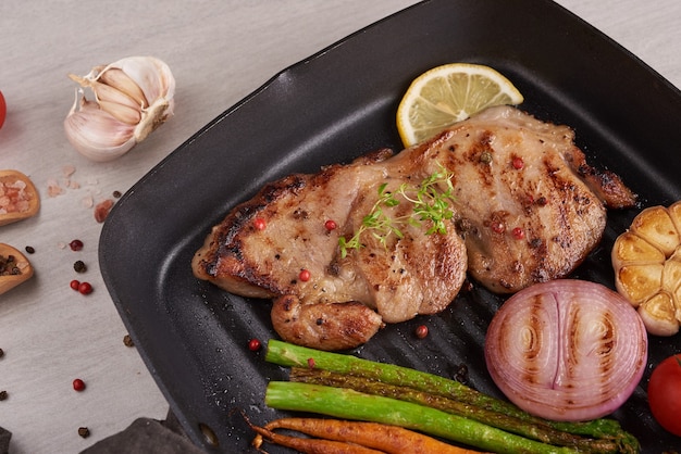 Gegrilltes Schweinesteak von einem Sommergrill mit Gemüse, Spargel, Babykarotten, frischen Tomaten und Gewürzen. Gegrilltes Steak auf Grillpfanne auf Steinoberfläche. Draufsicht.