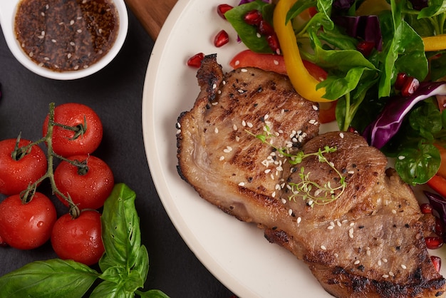 Gegrilltes Steak mit frischem Gemüse, Paprika, Tomaten, roten Zwiebeln, rosa Pfeffer und Gewürzen. Hausgemachtes leckeres Essen. Konzept für leckeres und gesundes Essen. Schwarze Steinoberfläche. Schweinesteak mit Salat