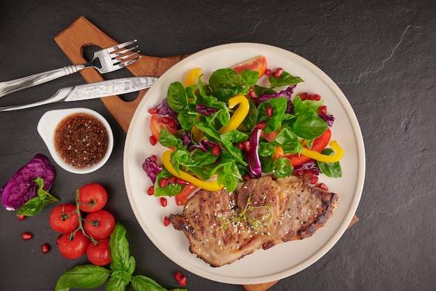 Gegrilltes Steak mit frischem Gemüse, Paprika, Tomaten, roten Zwiebeln, rosa Pfeffer und Gewürzen. Hausgemachtes leckeres Essen. Konzept für leckeres und gesundes Essen. Schwarze Steinoberfläche. Schweinesteak mit Salat