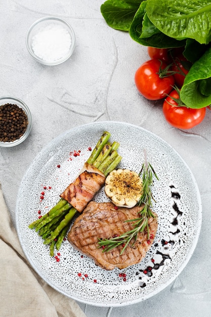 Gegrilltes Steak mit Spargel, Rosmarinblatt und gegrilltem Knoblauch auf Teller und Zutaten mit Gemüse auf Betontisch-Draufsicht