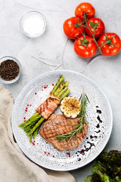 Gegrilltes Steak mit Spargel, Rosmarinblatt und gegrilltem Knoblauch auf Teller und Zutaten mit Gemüse auf Betontisch