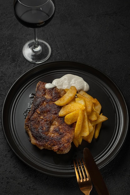 Gegrilltes Steak und Pommes Frites mit Sauce auf einem schwarzen Teller mit einem Glas Rotwein auf schwarzem Hintergrund