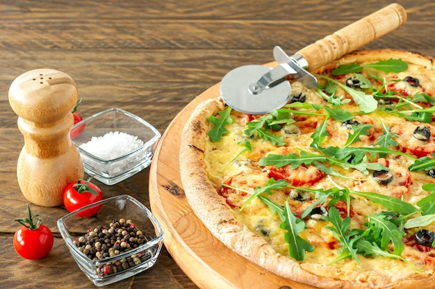 Gekochte heiße Pizzatorte mit Peperoni, Tomaten, schwarzen Oliven, Rucola und Mozzarella auf Holzbrett.