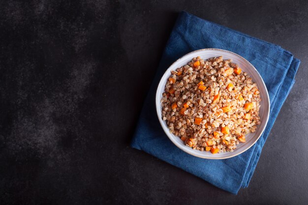 Gekochtes Buchweizen-Müsli mit Karotte, Petersilie und Butter in einer Schüssel auf dunklem Hintergrund. Traditionelles russisches Gericht auf dunklem Hintergrund mit Kopienraum