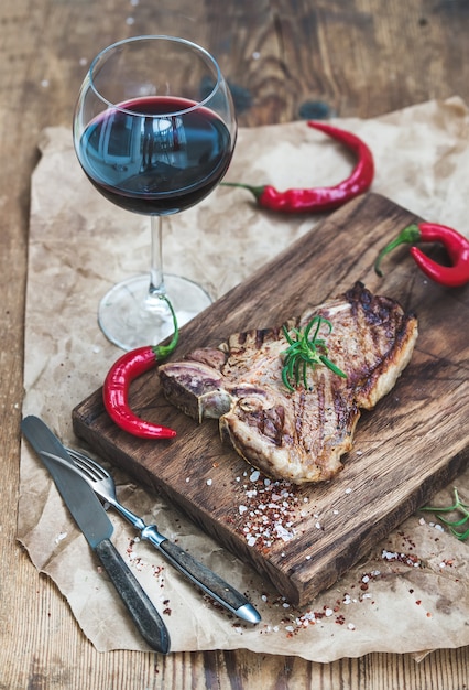 Gekochtes Fleischknochensteak auf Umhüllungsbrett mit gebratenen Tomaten, Paprikapfeffern, frischem Rosmarin, Gewürzen und Glas Rotwein über rustikaler Holzoberfläche