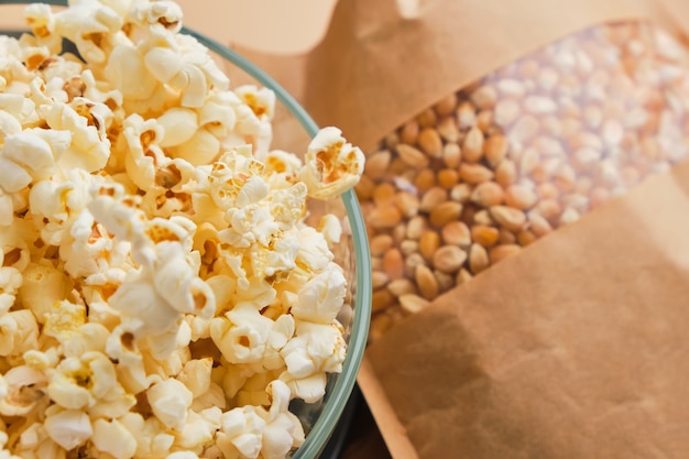 Gekochtes Popcorn in Glasschüssel und Maiskörner in einer Pappschachtel auf einer beigen Oberfläche