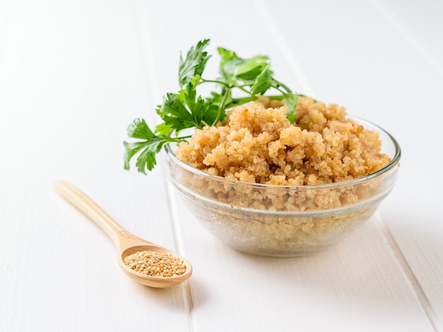 Gekochtes Quinoamüsli in einer Glasschüssel auf einem weißen Holztisch mit Petersilienblättern.