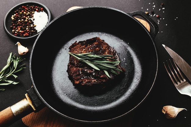 Gekochtes Ribeye-Steak auf gusseiserner Pfanne und Brett mit Pfeffer, Rosmarin, Salz, Knoblauch, Messer, Gabel, Butter auf schwarzem Hintergrund