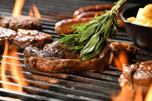 Gekochtes, saftiges Steakfleisch mit Rosmarin bestrichenes Rindfleisch auf einem flammenden Grill, umgeben von anderem Fleisch und Würstchen