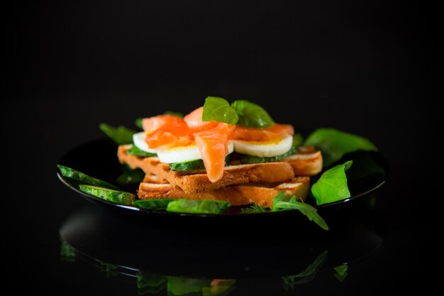 Gekochtes Sandwich mit leicht gesalzenen roten Fischgurken und gekochten Eiern mit Kräutern isoliert auf schwarzem Hintergrund