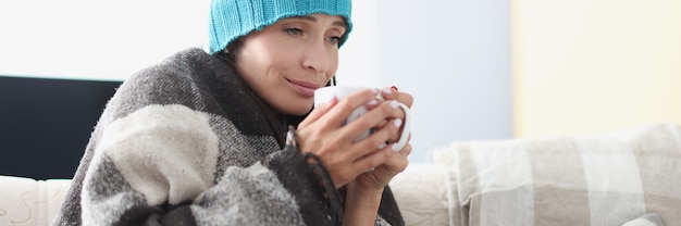 Gekühlte Frau auf der Couch in Decke und Hut hält heiße Tasse