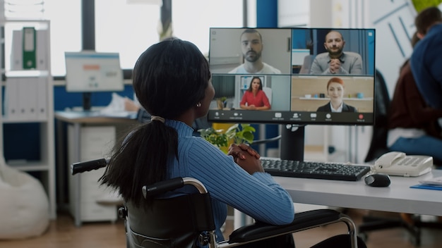 Gelähmter behinderter schwarzer Arbeiter, der im Rollstuhl immobilisiert sitzt und ein Videomeeting hat, das online mit Kollegen aus der Ferne im Geschäftsbüro diskutiert discuss