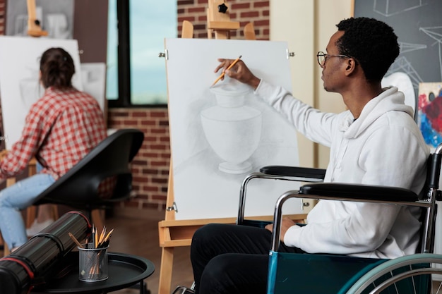 Gelähmter Schüler im Rollstuhl, der am Kunstunterricht teilnimmt und Vasenskizze auf weißer Leinwand mit grafischem Bleistift zeichnet, der an der Skizziertechnik arbeitet. Vielfältiges Team, das Draw-Fähigkeiten für persönliches Wachstum entwickelt