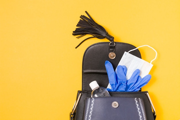 Gelalkohol-Desinfektionsmittel, Gesichtsmaske und Einweghandschuhe aus blauem Gummi in einer Handtasche auf gelbem Grund