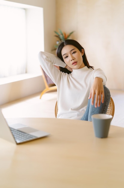 Gelangweilte junge asiatische Frau in warmer Pulloverkleidung, die mit Laptop im Heimbüro am Tisch sitzt, hat ein Problem mit der Fernarbeit. Fühlen Sie sich deprimiert, müde, faul während der Arbeit im Heimbüro