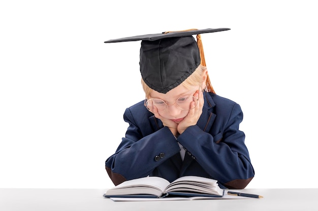 Gelangweilter Junge in Schuluniform und Studentenhut sitzt am Tisch und liest Lehrbuch Das Kind hat es satt zu lernen Der Schuljunge langweilt sich