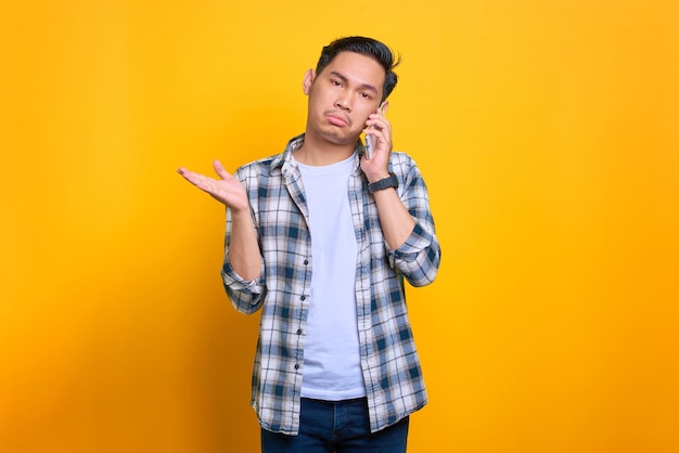 Gelangweilter junger asiatischer Mann im karierten Hemd, der am Handy spricht, isoliert auf gelbem Hintergrund
