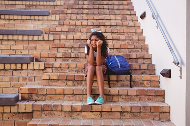 Gelangweiltes afrikanisch-amerikanisches Schulmädchen sitzt mit Schultasche auf Stufen im Schulhof