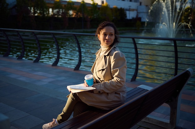 Gelassene Frau in beigem Trenchcoat, die auf einer Parkbank auf dem Seehintergrund sitzt, Kaffee oder heißes Getränk in recycelbarem Pappbecher zum Mitnehmen trinkt und ein Buch liest und sich von digitalen Geräten ausruht