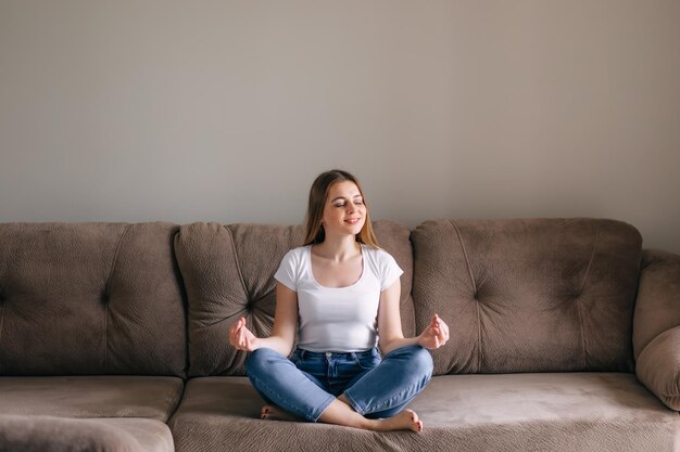 Gelassenes, gesundes Mädchen, das mit geschlossenen Augen meditiert, sitzt zu Hause auf dem Sofa in Lotusposition, achtsame junge Frau, die sich beim Yoga entspannt