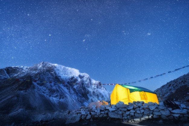 Gelb leuchtendes Zelt gegen hohe Felsen mit schneebedecktem Gipfel und Himmel mit Sternen