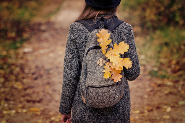 Gelbe Blätter in einem Rucksack, ein Mädchen steht in einem herbstlichen Wald. Rückansicht