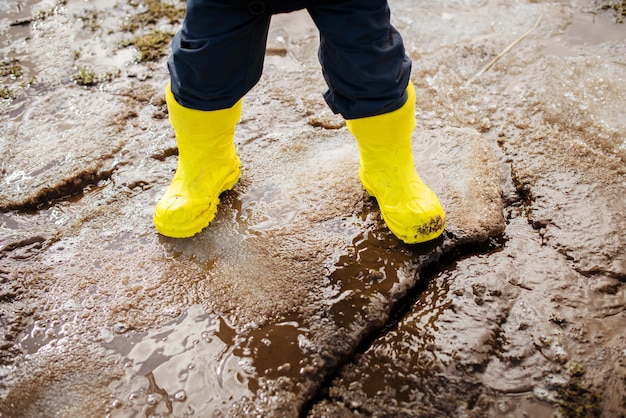 gelbe Gummistiefel an den Füßen eines Jungen, der im zeitigen Frühjahr durch Schlammschnee und Pfützen geht