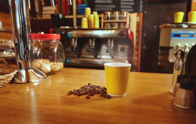 Gelbe pappbecher mit heißem kaffee und verstreuten gerösteten bohnen auf einem holztisch auf dem hintergrund der kaffeemaschine