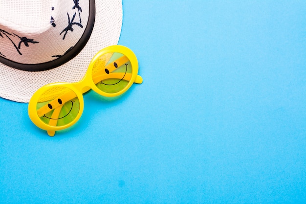 Gelbe Plastiksonnenbrille mit gemalten Augen und einem Lächeln auf den Gläsern und einem Hut auf einem blauen Hintergrund