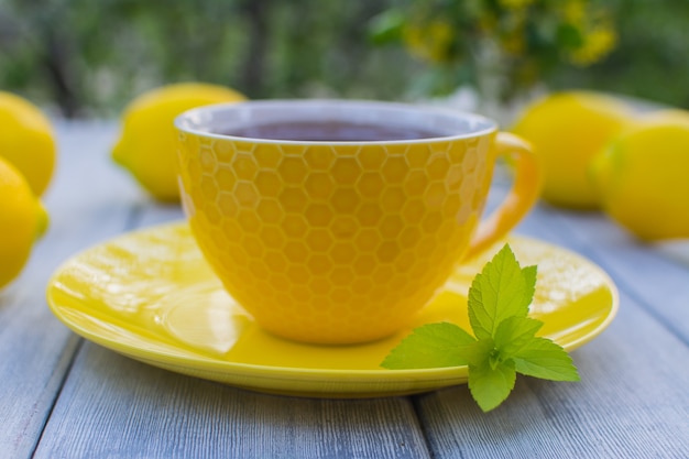 Gelbe Porzellantasse mit Tee mit gelber Untertasse auf einem Holztisch Im Hintergrund sind gelbe Zitronen