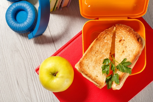 Gelbe Sandwichbox mit gerösteten Brotscheiben, Käse und grüner Petersilie, grünem Apfel, Kopfhörern und gebundenen Büchern im Hintergrund. Schulfrühstück.