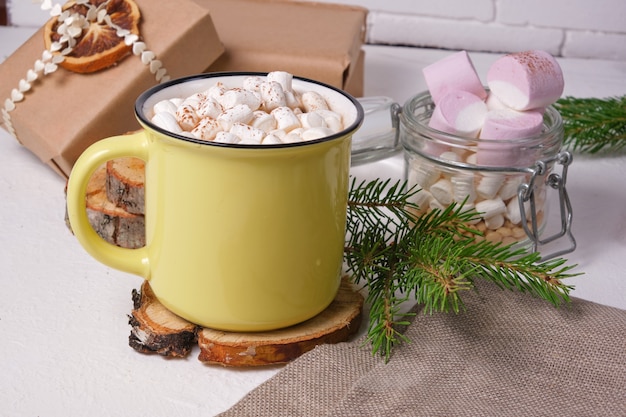 Gelbe Tasse mit Kakao verziert mit Marshmallows auf einem Ständer aus Sägeschnittholz, einem Glas Marshmallows, einem Fichtenzweig und Weihnachtsgeschenken im Hintergrund
