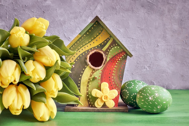 Gelbe Tulpen, Vogelhäuschen und bemalte Ostereier mit dekorativer Filzblume
