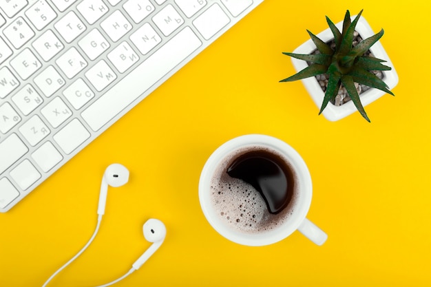 Gelber Arbeitsplatz mit Tastatur, Kopfhörern, Tasse schwarzen Kaffees und Blumentopf.