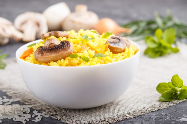 Gelber gebratener Reis mit Champignons pilzt Gelbwurz und Oregano in der weißen keramischen Schüssel auf einem schwarzen konkreten Hintergrund