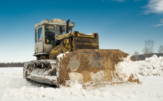 Gelber Raupentraktor-Bulldozer mit einem Eimer arbeitet im Winter und räumt die Straße vom Schnee ab.