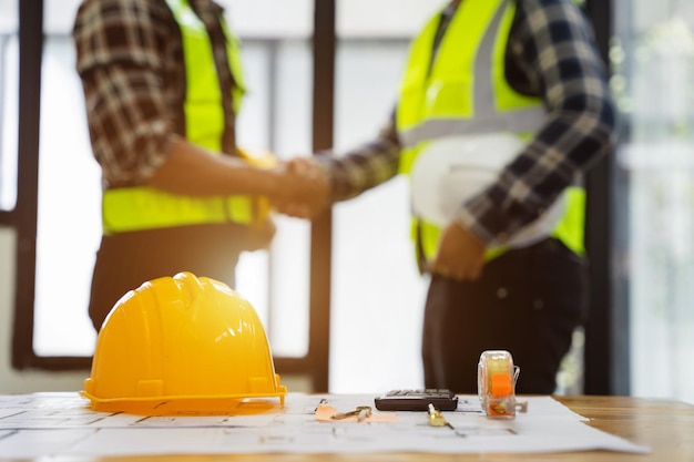 Gelber Schutzhelm auf Werkbank mit Bauarbeiterhänden, die Begrüßungsstartplan schütteln, um neues Projekt im Baubürozentrum und Bauunternehmerkonzept abzuschließen