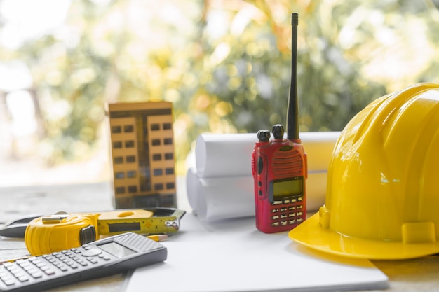 gelber Sicherheitshelm und aufgerollte architektonische Baupläne auf einem Holzschreibtisch, die auf einen Arbeitsbereich eines Architekten hindeuten, möglicherweise auf einer Baustelle