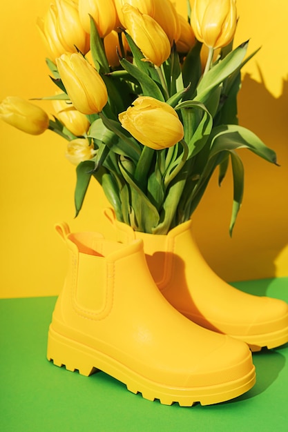 Gelber Tulpenblumenstrauß in gelben Gummistiefeln als Vase auf gelbem und grünem Hintergrund mit scharfen Schatten Kreative Frühlingsblumenkomposition Botaniktapete Wohnkultur