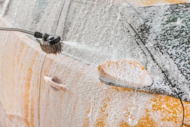 Gelbes Auto wird in Selbstbedienungswaschanlage gewaschen. Shampoo und Schaum sprühen auf Seitenspiegel und Fenster.