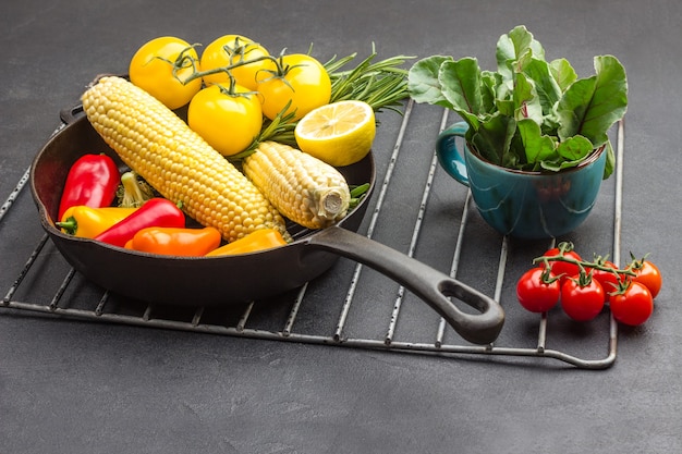 Gelbes Gemüse in der Pfanne, Gemüse im Becher und ein Zweig roter Tomaten auf dem Metallgrill.