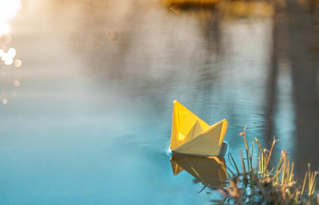 Gelbes schwimmendes Papierboot Buntes Segelschiff in großer blauer Frühlingspfütze Flusswasser auf der Winterstraße Warmes nasses Regenwetter altes Gras Hallo Frühling Herbst Kinder spielen viel Spaß im Freien