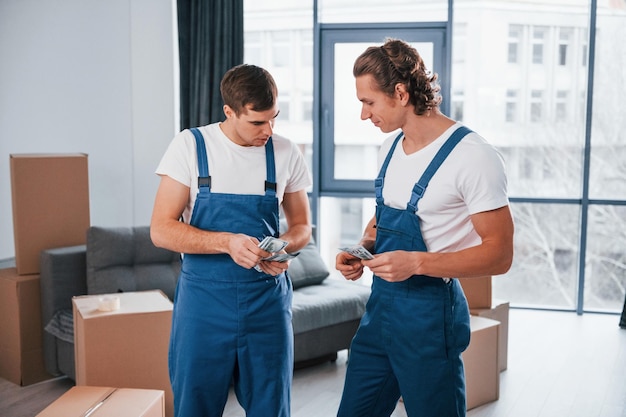 Geld halten Zwei junge Mover in blauer Uniform, die drinnen im Raum arbeiten