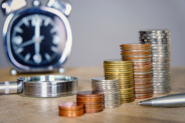 Geldmünzen, die mit Uhr- und Vergrößerungsglasglas für das Spar- und Investitionskonzept stapeln.