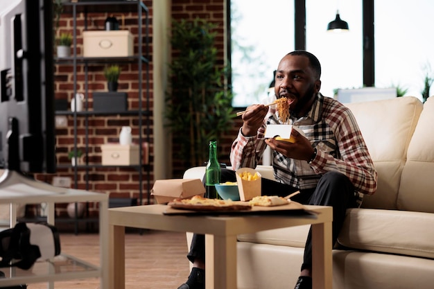 Gelegenheitsperson, die Nudelgerichte aus der Lieferbox genießt und einen Film im Fernsehprogramm ansieht. Essen zum Mitnehmen mit Stäbchen zur Unterhaltung, Flasche Bier.