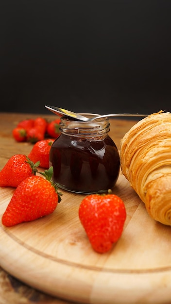 Geléia de morango na jarra de vidro com colher de metal com frutas frescas e croissant em fundo de madeira ingredientes caseiros para fazer café da manhã