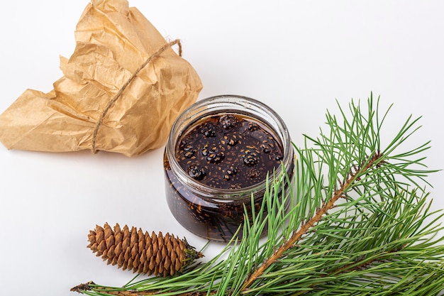 Geléia incomum de pinhas em frasco de vidro em fundo branco entre galhos de pinheiro e cones. Fim doce orgânico e da sobremesa do vegetariano acima, copie o espaço para o texto. Remédio para resfriados